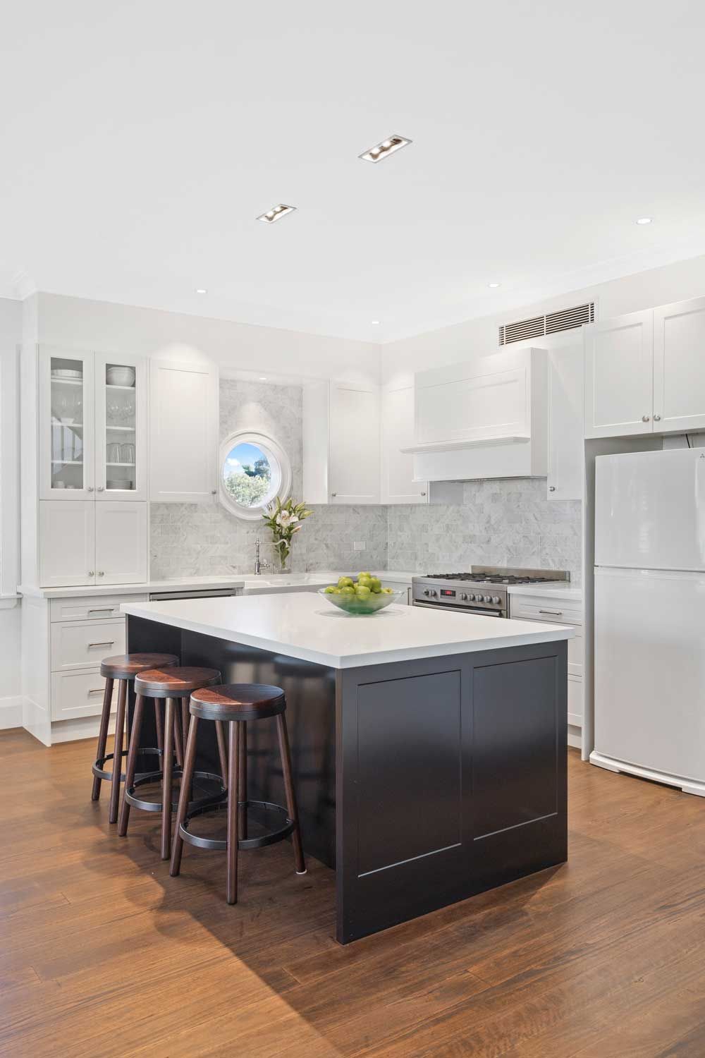 Kitchen renovation with black island, white shaker cabinets, marble backsplash, porthole window, and timber floors.