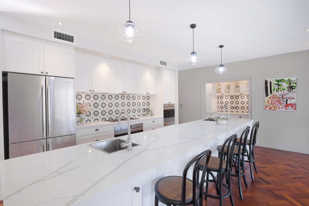 Modern kitchen renovation with white shaker cabinets, marble waterfall island, patterned tile backsplash, and stainless steel fridge.