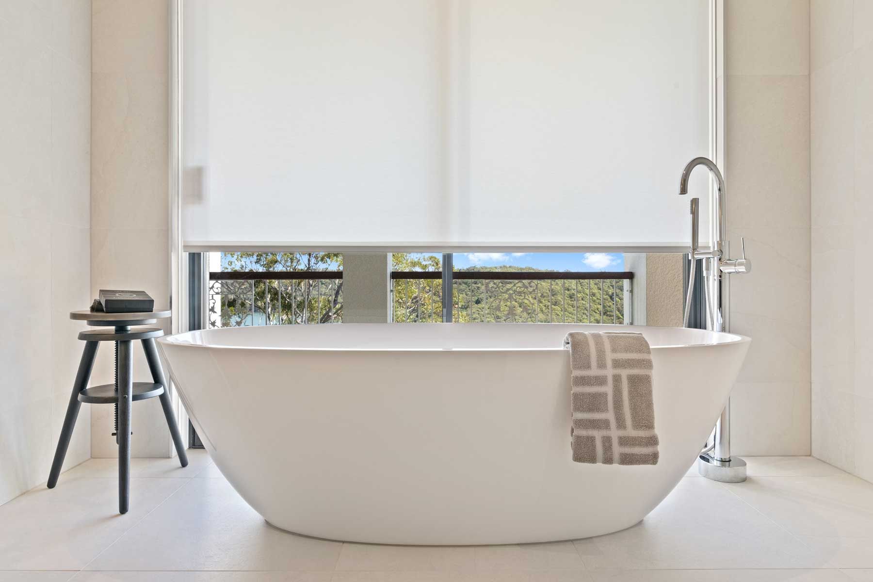 Modern bathroom renovation with white freestanding oval bathtub, floor-mounted chrome mixer tap, and white roller blind.