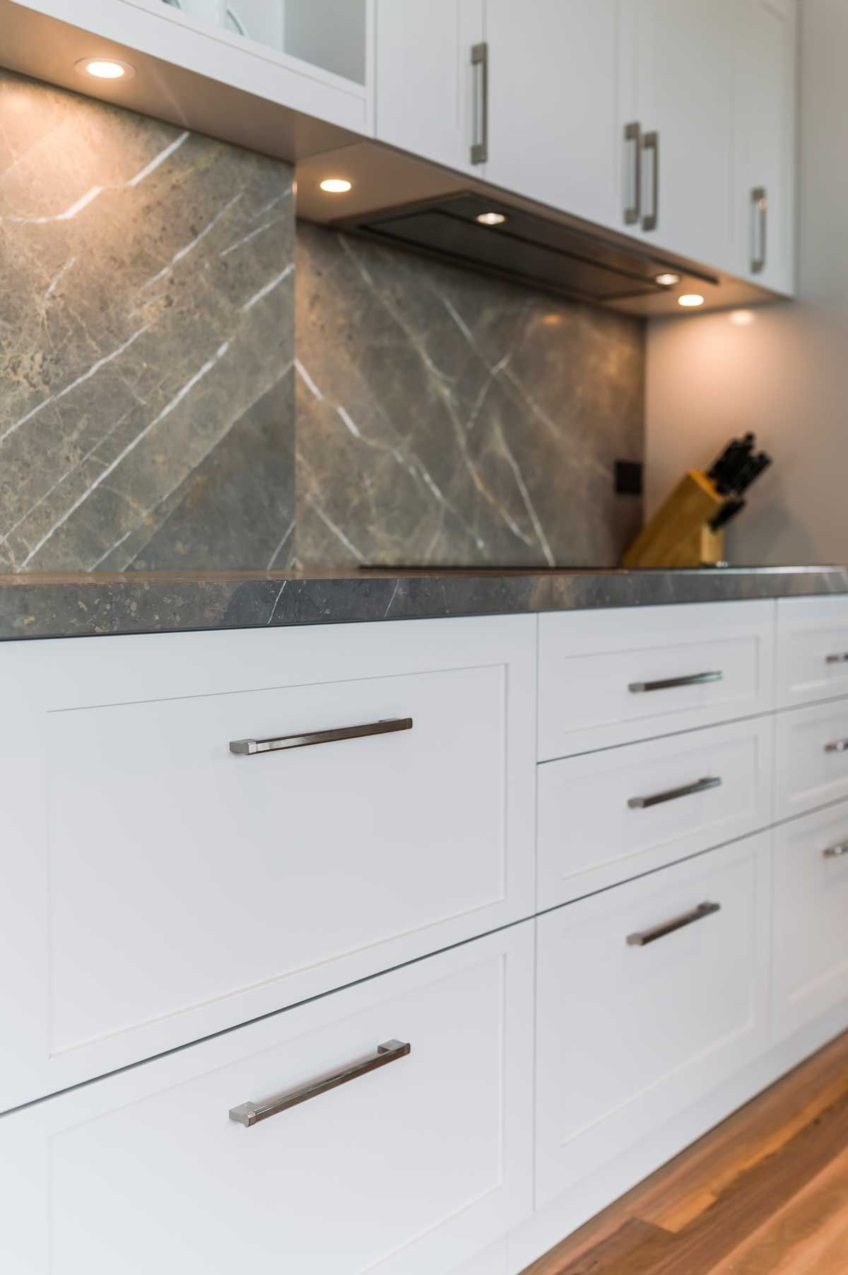 Modern kitchen renovation with white shaker drawers, gray marble backsplash, dark stone benchtop, and under-cabinet LED lighting.