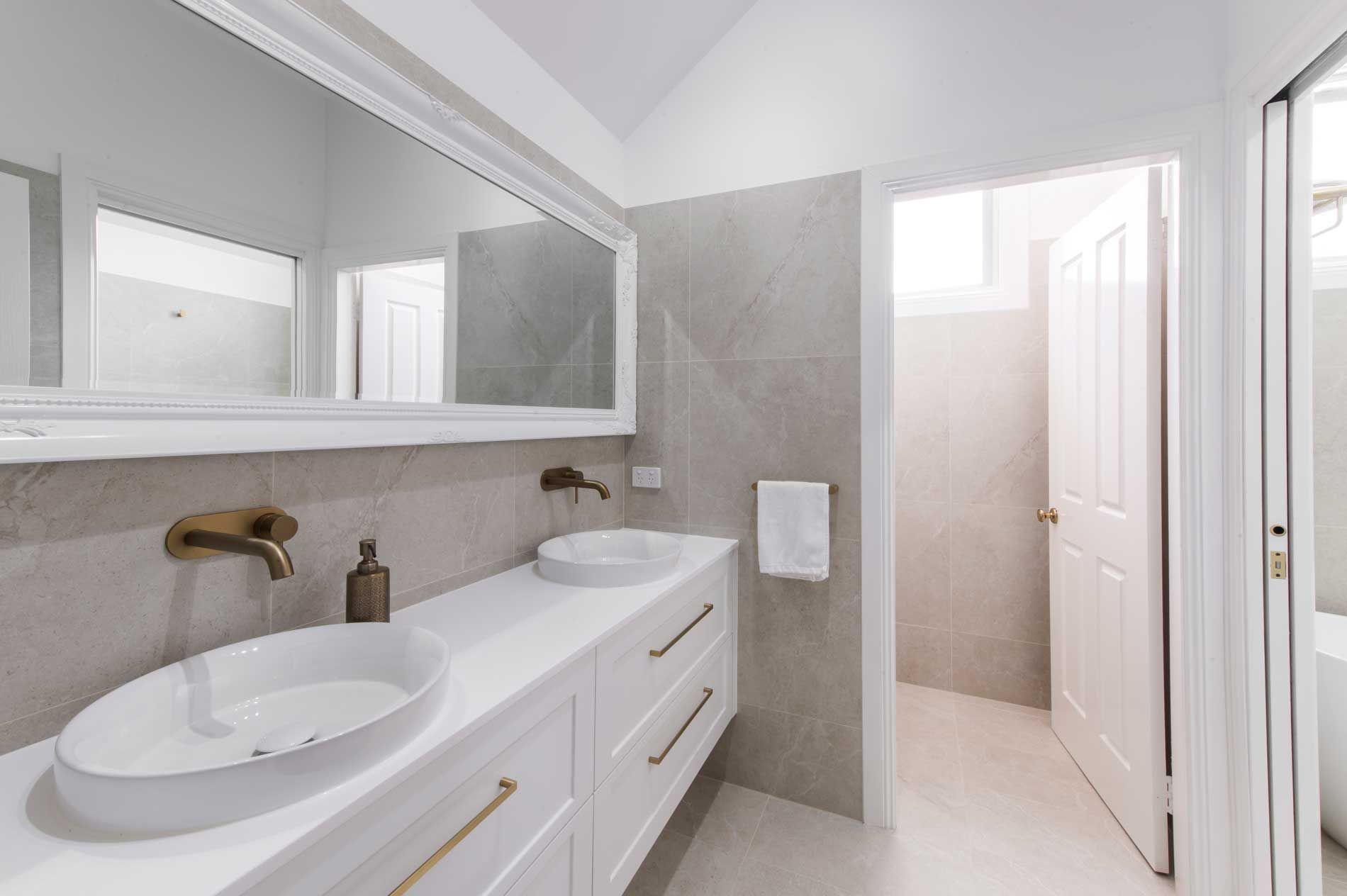 Modern bathroom renovation with white shaker double vanity, brushed gold wall-mounted taps, and large white-framed mirror.