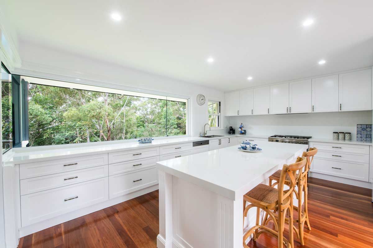 Contemporary kitchen renovation featuring a large white island with timber stools, deep storage drawers, and a wide window splashback.