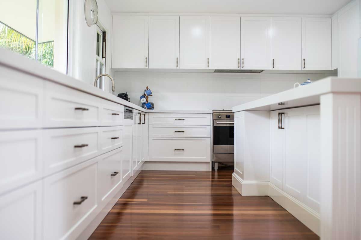 Modern white kitchen renovation with shaker-style cabinets, black handles, white stone benchtops, and polished timber floors.