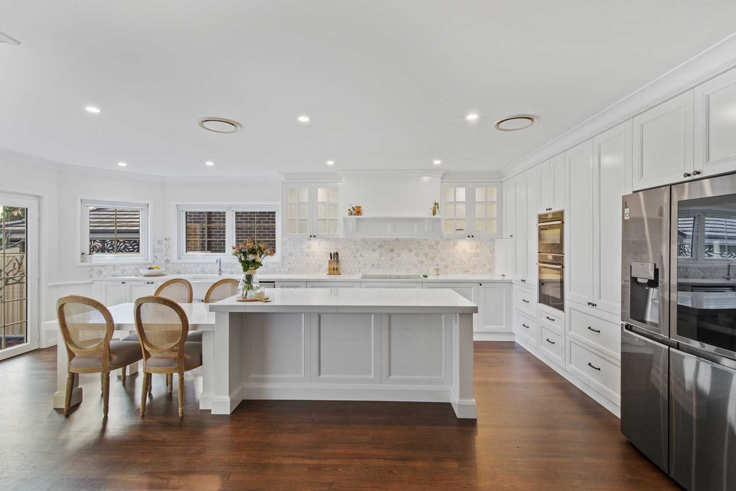 Hamptons-style kitchen renovation with white shaker cabinets, marble hexagonal tile backsplash, white island, and dark timber floors.