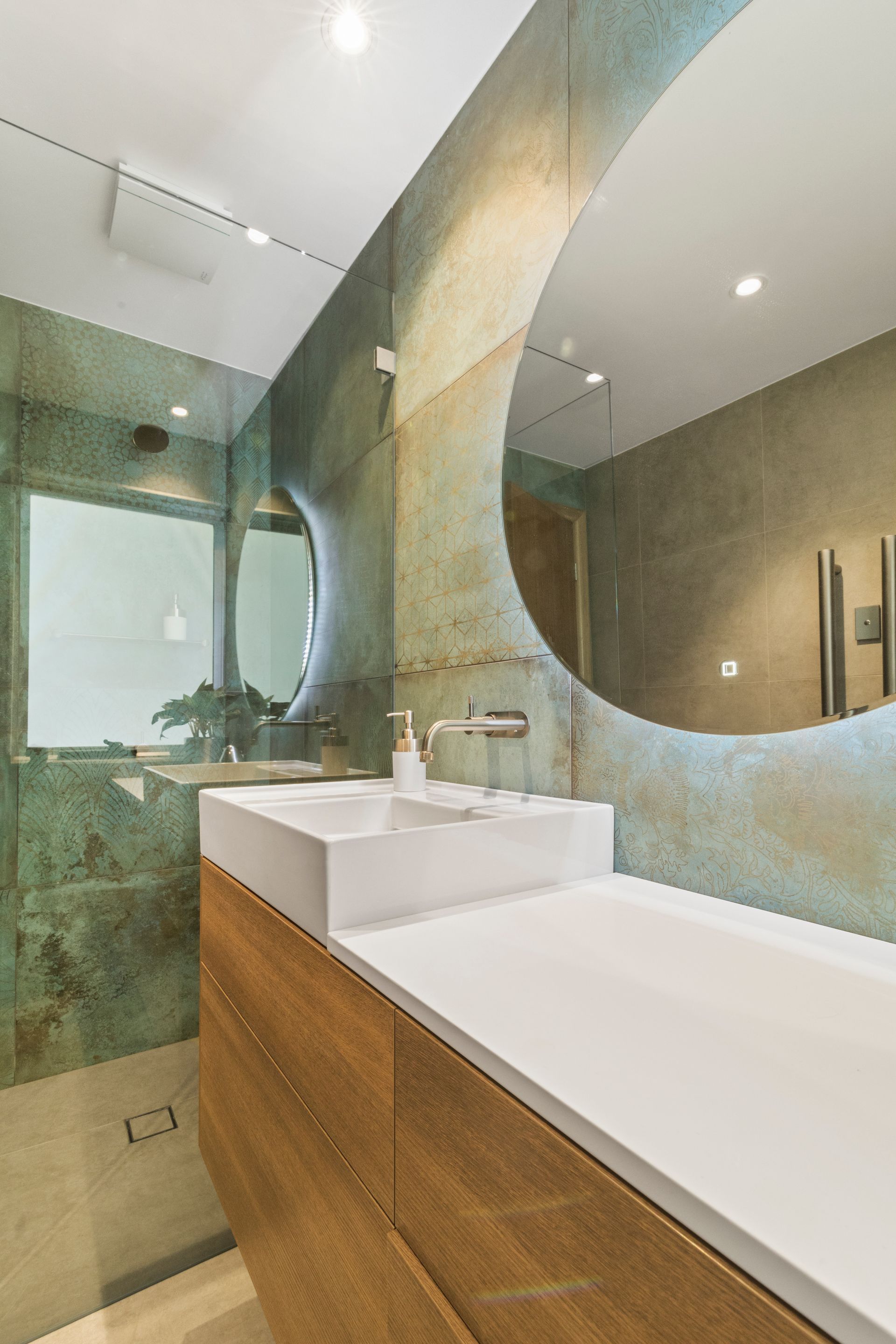 Modern bathroom renovation with oxidized copper-look wall tiles, a backlit circular mirror, white vessel sink, and timber floating vanity.