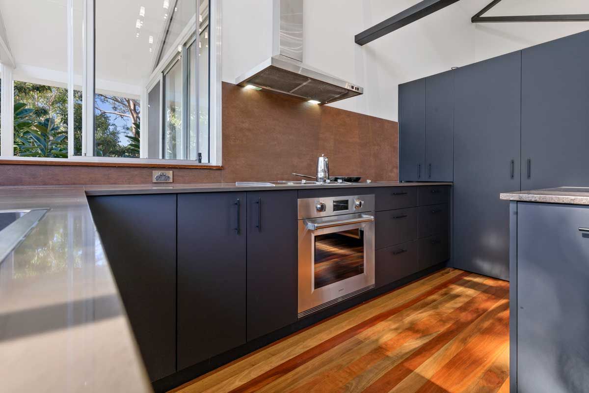 Modern kitchen renovation with matte charcoal cabinetry, stainless steel oven, copper-tone backsplash, and polished timber floors.