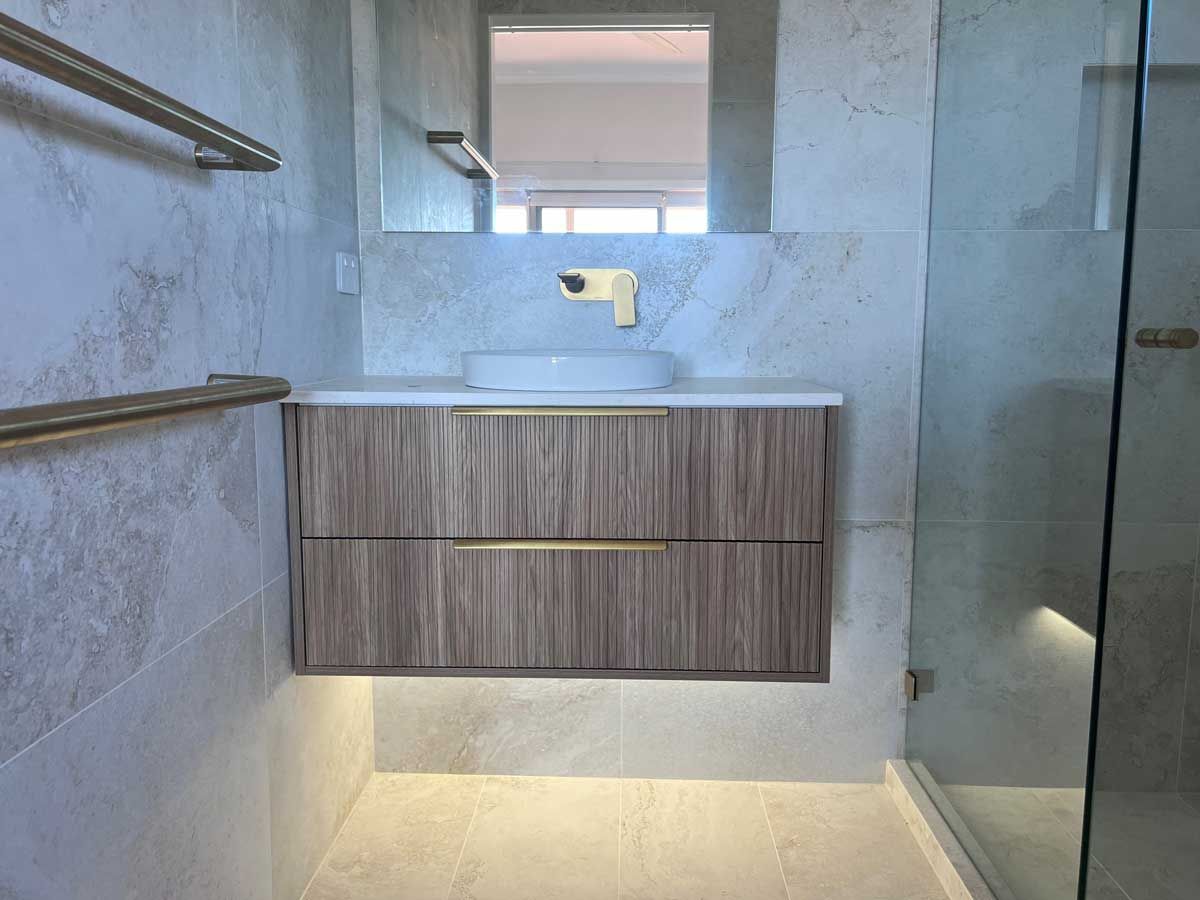 Contemporary floating wood vanity with gold hardware, backlit by LED strips, against gray marble-look wall tiles.