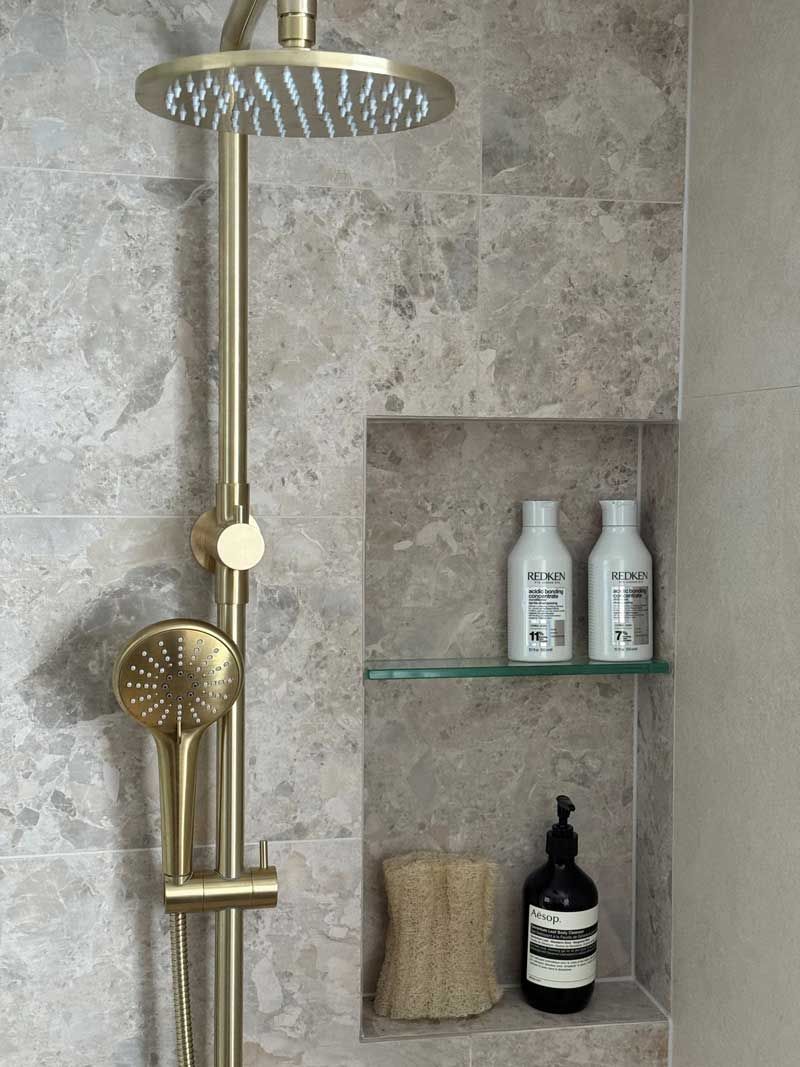 Modern shower renovation with brushed gold dual shower head system, gray marble-look wall tiles, and a recessed tiled niche.