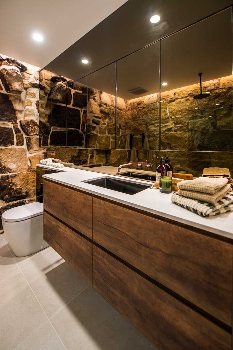 Luxury master bathroom featuring an exposed sandstone wall, integrated white countertop sink, and minimalist wood cabinetry.