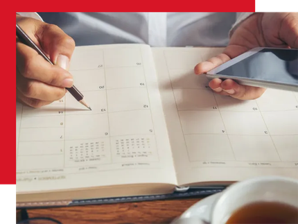 A person holds a smartphone in one hand and uses a pencil to write in a paper planner at a desk.