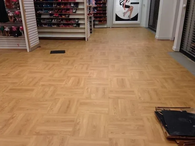 Interior of a shoe store with light brown parquet flooring. Shelves of shoes and a poster are visible.