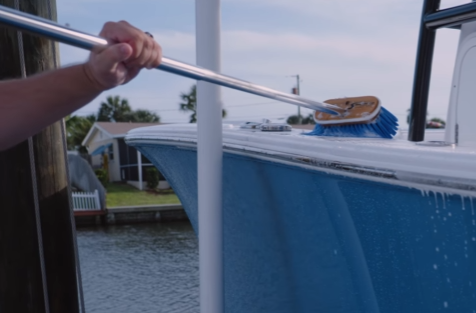 With a deck brush scrubbing white skid deck of a blue boat on a dock lift.