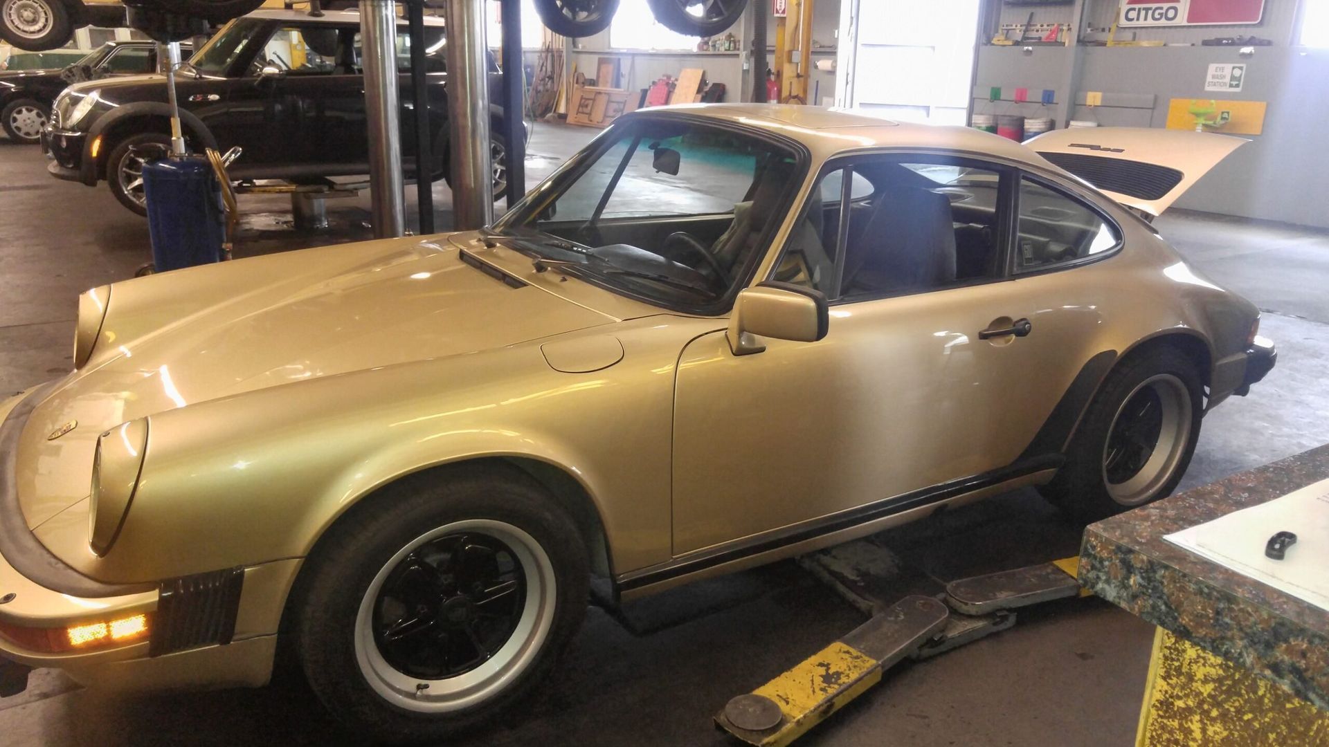 Gold Porsche 911 in a repair shop with its hood open, near a lift.