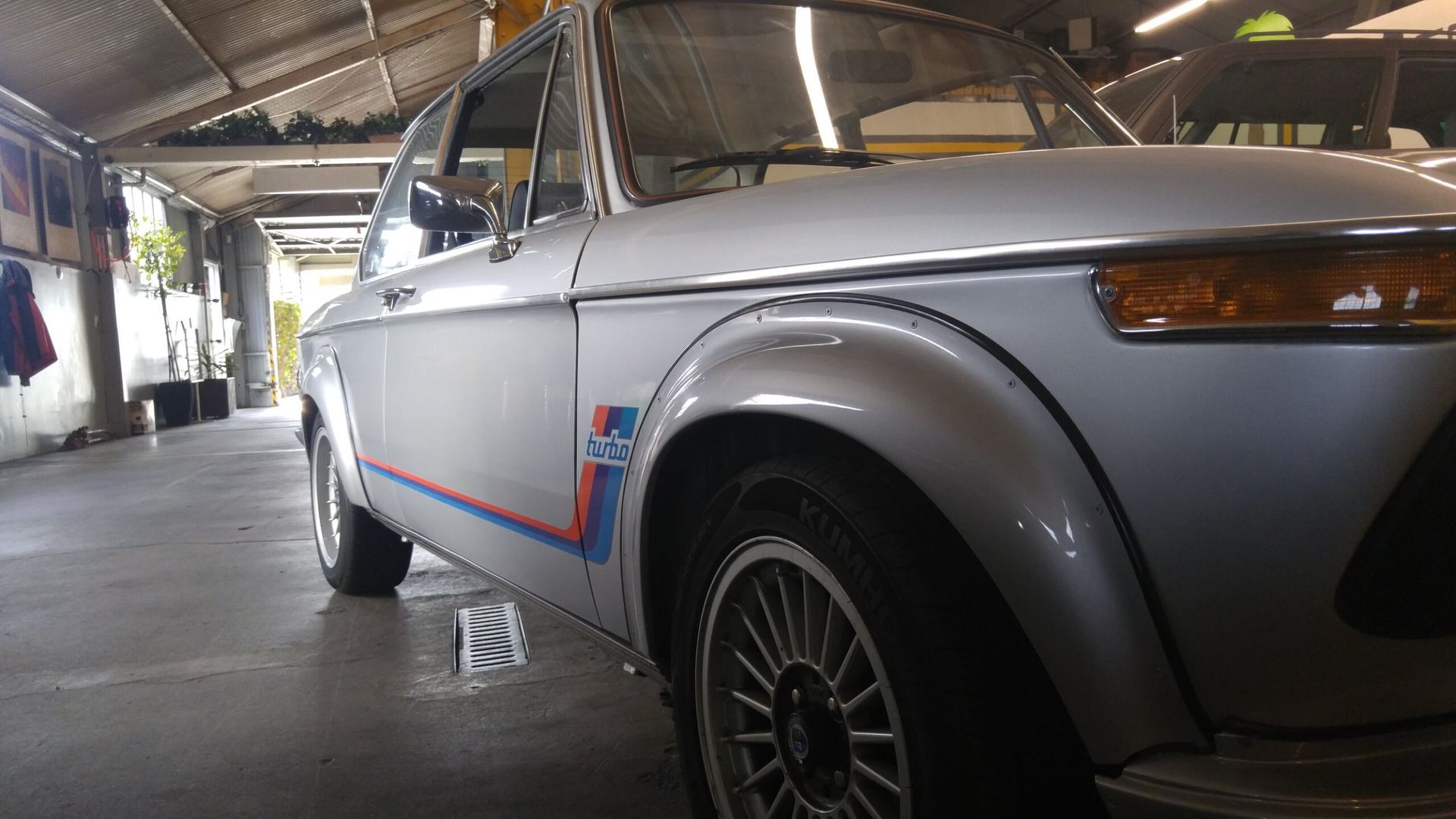 Silver vintage BMW 2002 with blue/red stripe detail, parked inside a garage.