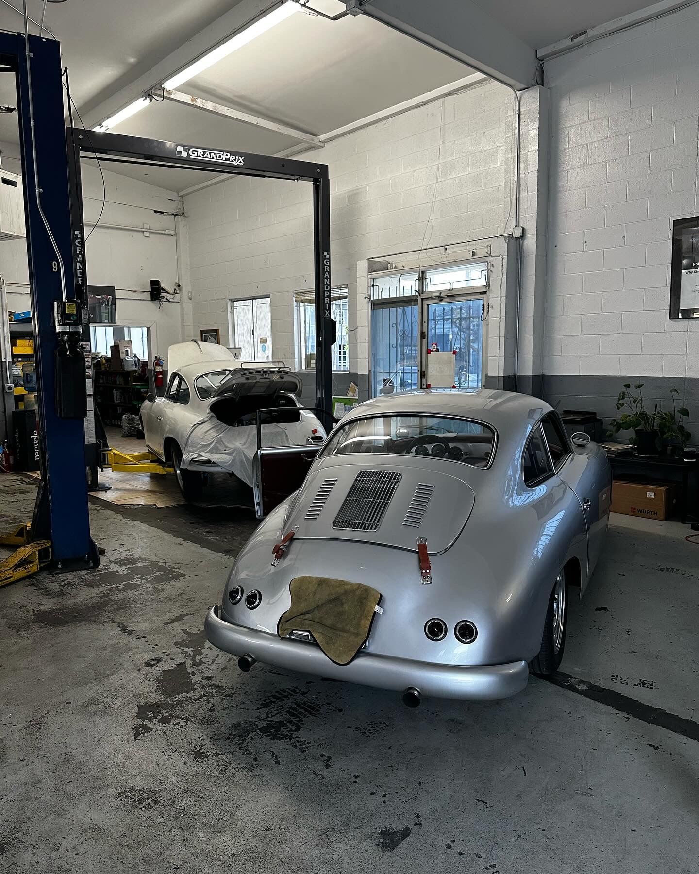 Silver vintage Porsche in a garage next to another car, both under lifts.