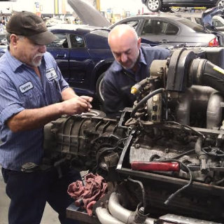 Two mechanics working on a car engine in a garage. One wears a cap and the other is bald.