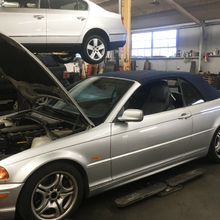 Silver convertible car in a repair shop with its hood open, another car lifted above it.
