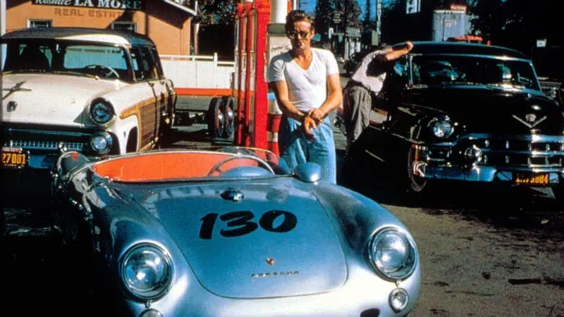 James Dean posing with his silver Porsche 550 Spyder, two cars, gas station backdrop.