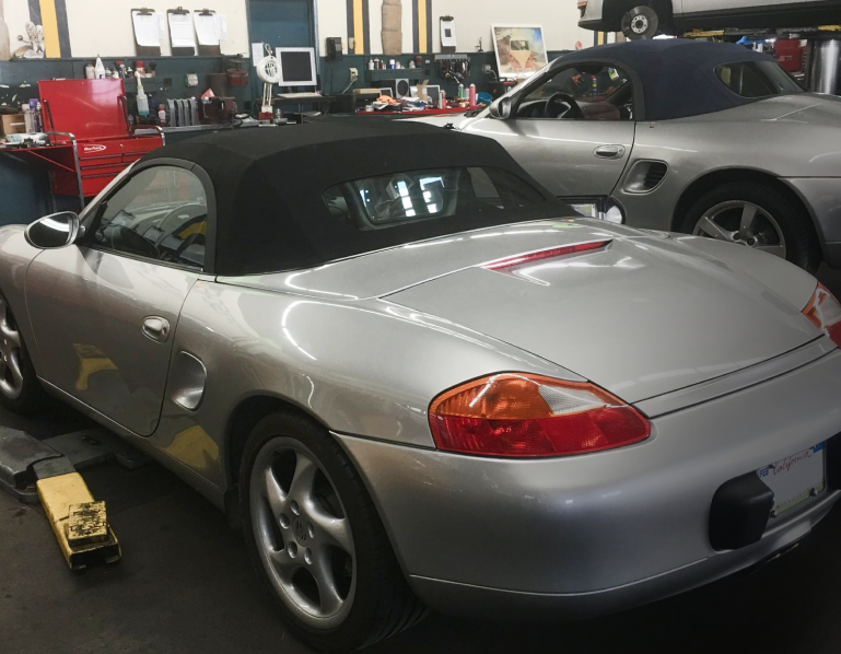 Silver Porsche Boxster convertible in a repair shop. Black top, another car in the background.