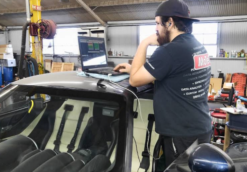 Mechanic with laptop on car hood, appears to be programming. In a garage setting.