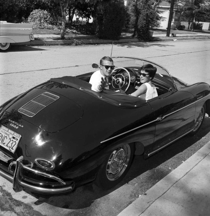 Man and woman in sunglasses driving a black vintage convertible Porsche on a sunny street.