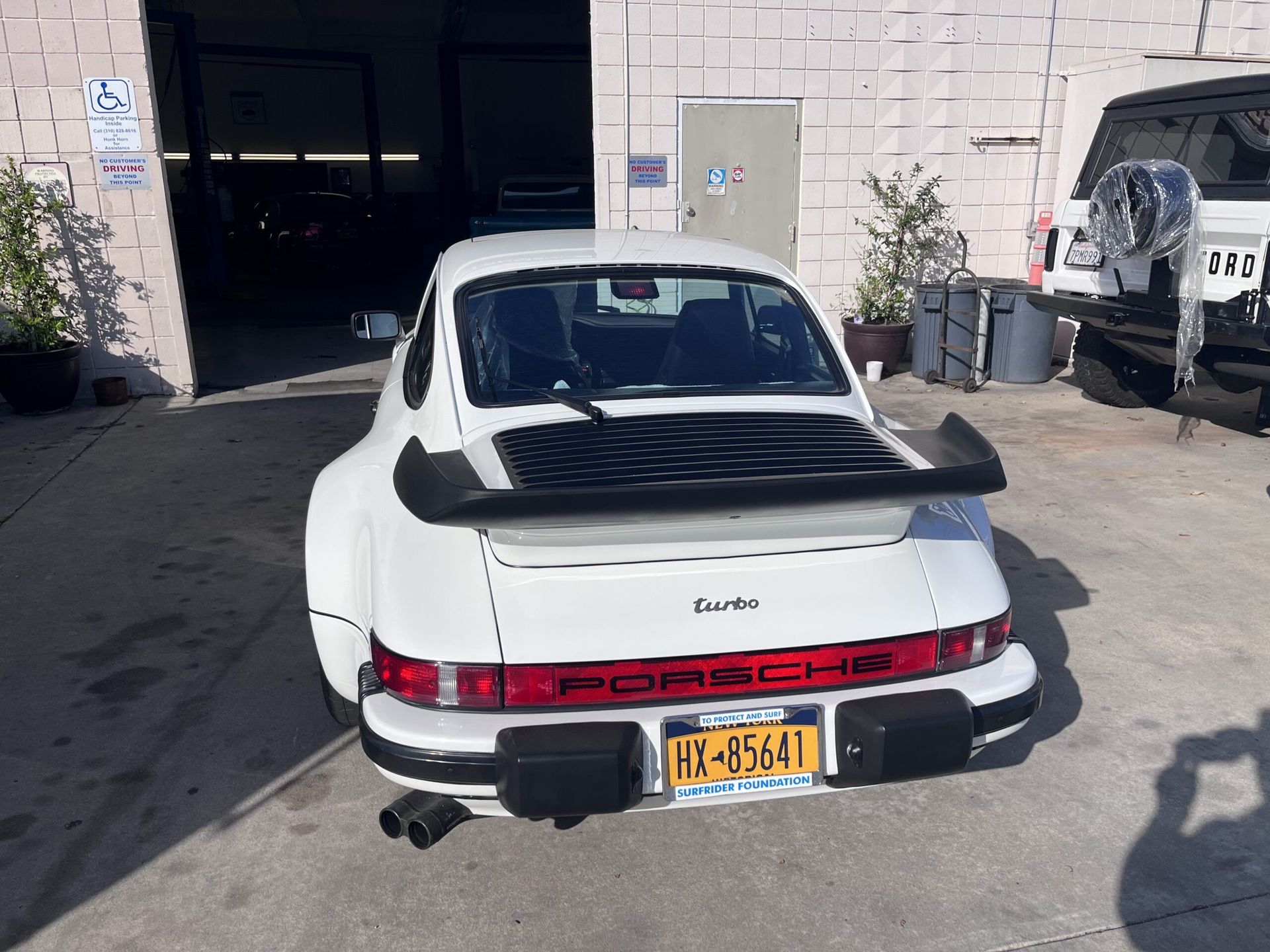 White Porsche Turbo sports car, parked outside garage. Rear view showing spoiler, New York license plate.