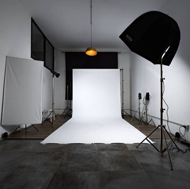 Una persona si trova in piedi su uno sfondo bianco in studio, di fronte a una troupe cinematografica che la sta riprendendo in un magazzino scarsamente illuminato.