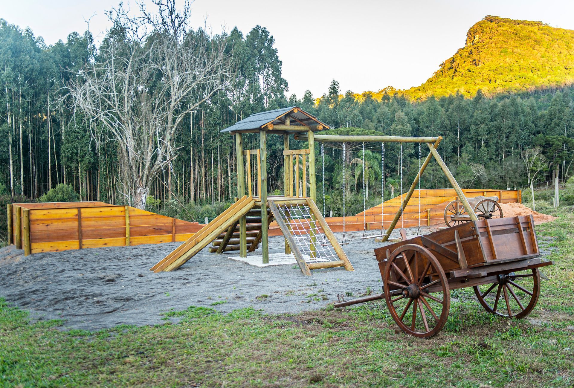 Um playground de madeira com um carrinho em primeiro plano e uma montanha ao fundo.