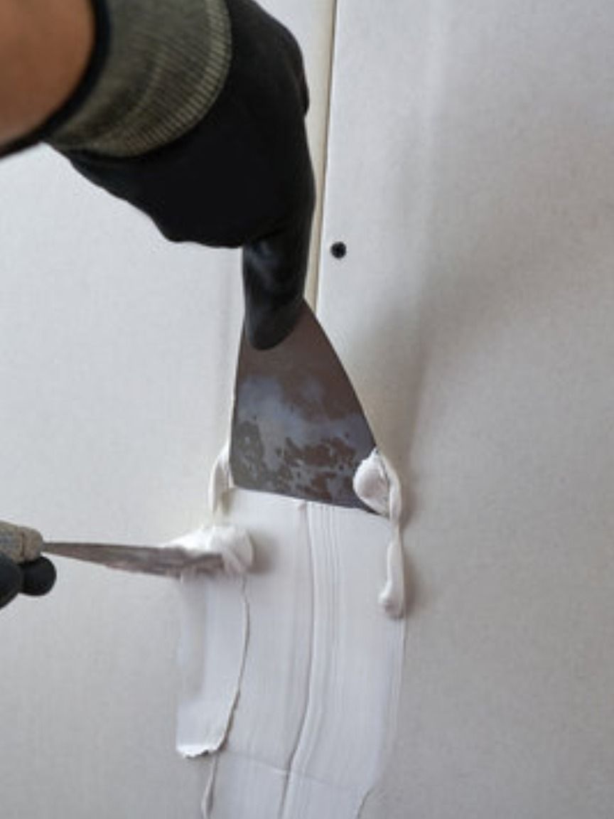 Person smoothing white joint compound between drywall panels with a metal trowel, wearing black gloves.