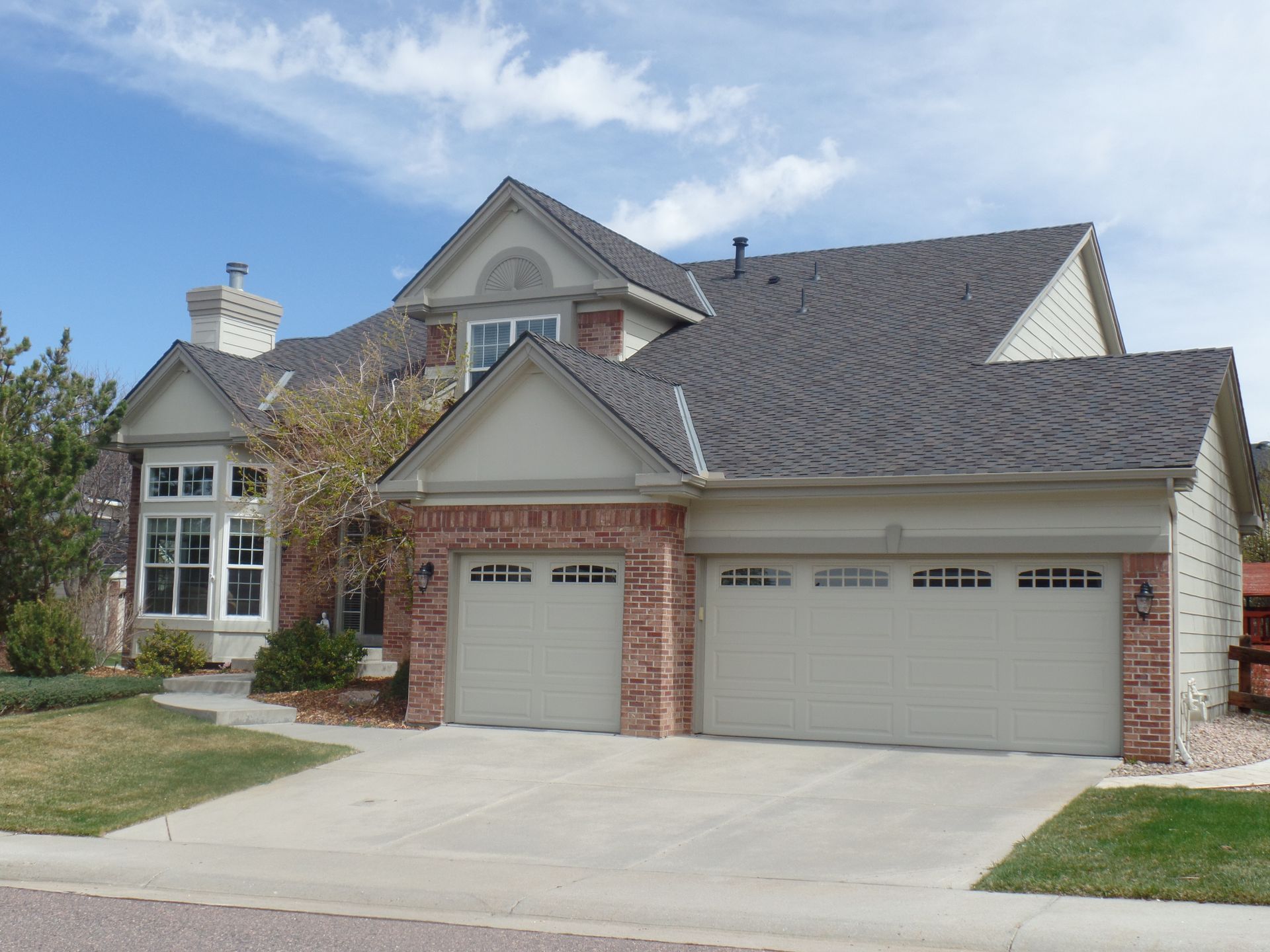 A two-story suburban house with brick and tan siding, a three-car garage, and a concrete driveway under a blue sky.
