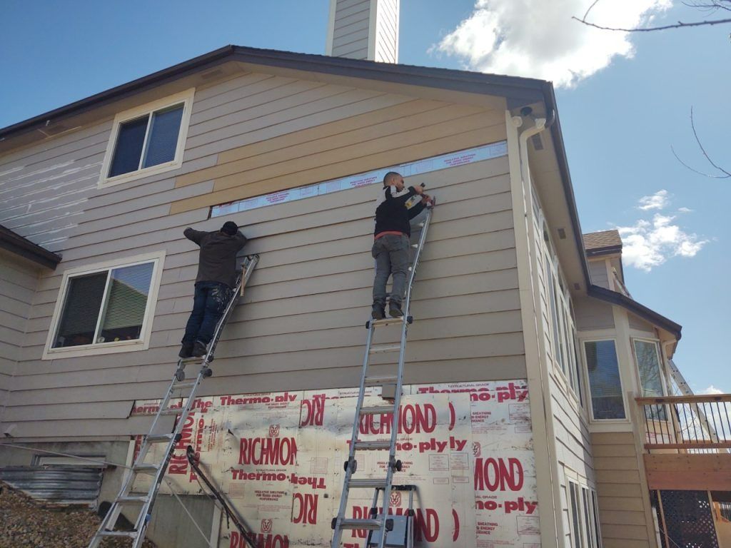 Two people on ladders installing siding on a house, sunny day.