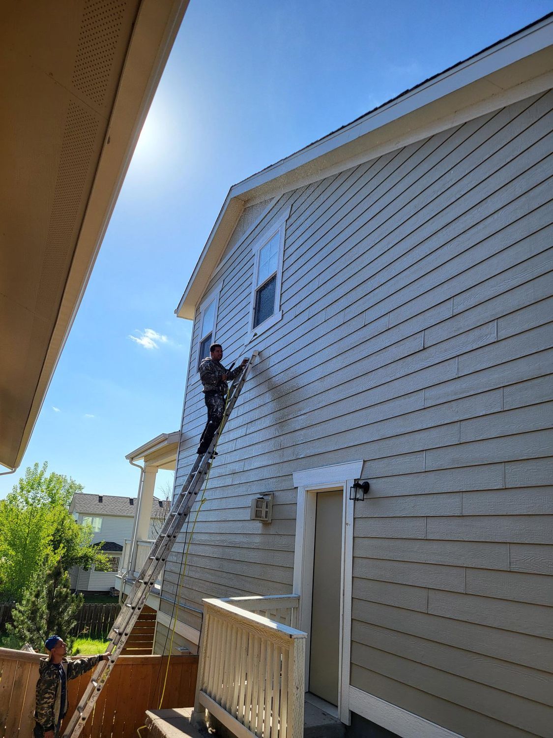 A person on a tall ladder uses a power washer to clean the light-colored siding of a house on a sunny day.