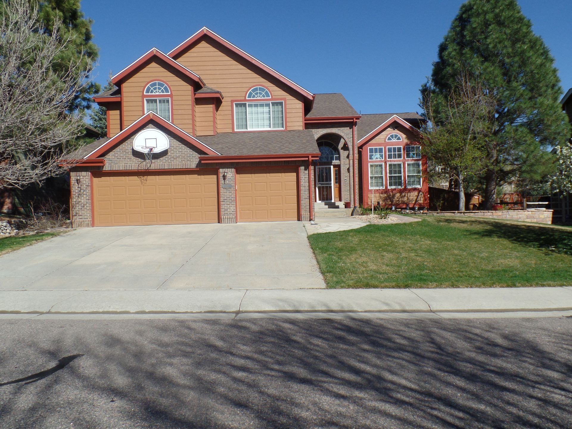 A two-story, tan suburban home with a two-car garage, basketball hoop, and driveway on a sunny day.