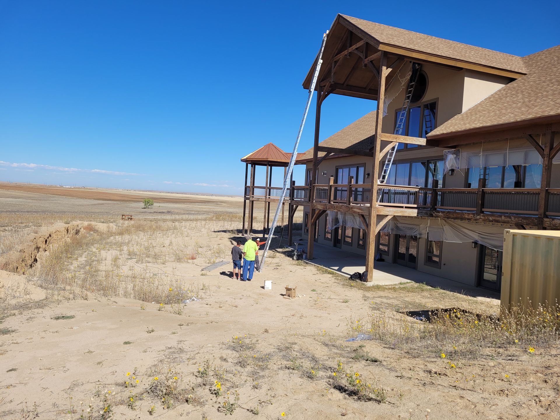 Two people work near a large, multi-story home with a balcony and a tall ladder, set in a vast, sandy, open landscape.