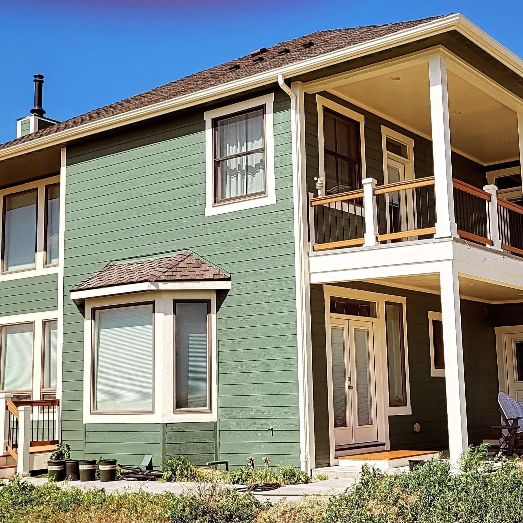 A two-story house with sage green shingle siding, white trim, a wrap-around porch, and a large bay window.