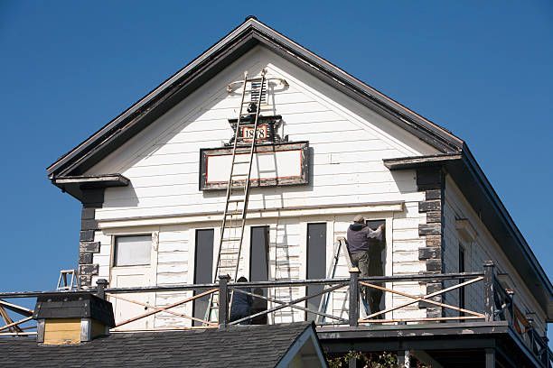 Workers use ladders to perform repairs on the exterior of a white, gabled house against a clear blue sky.