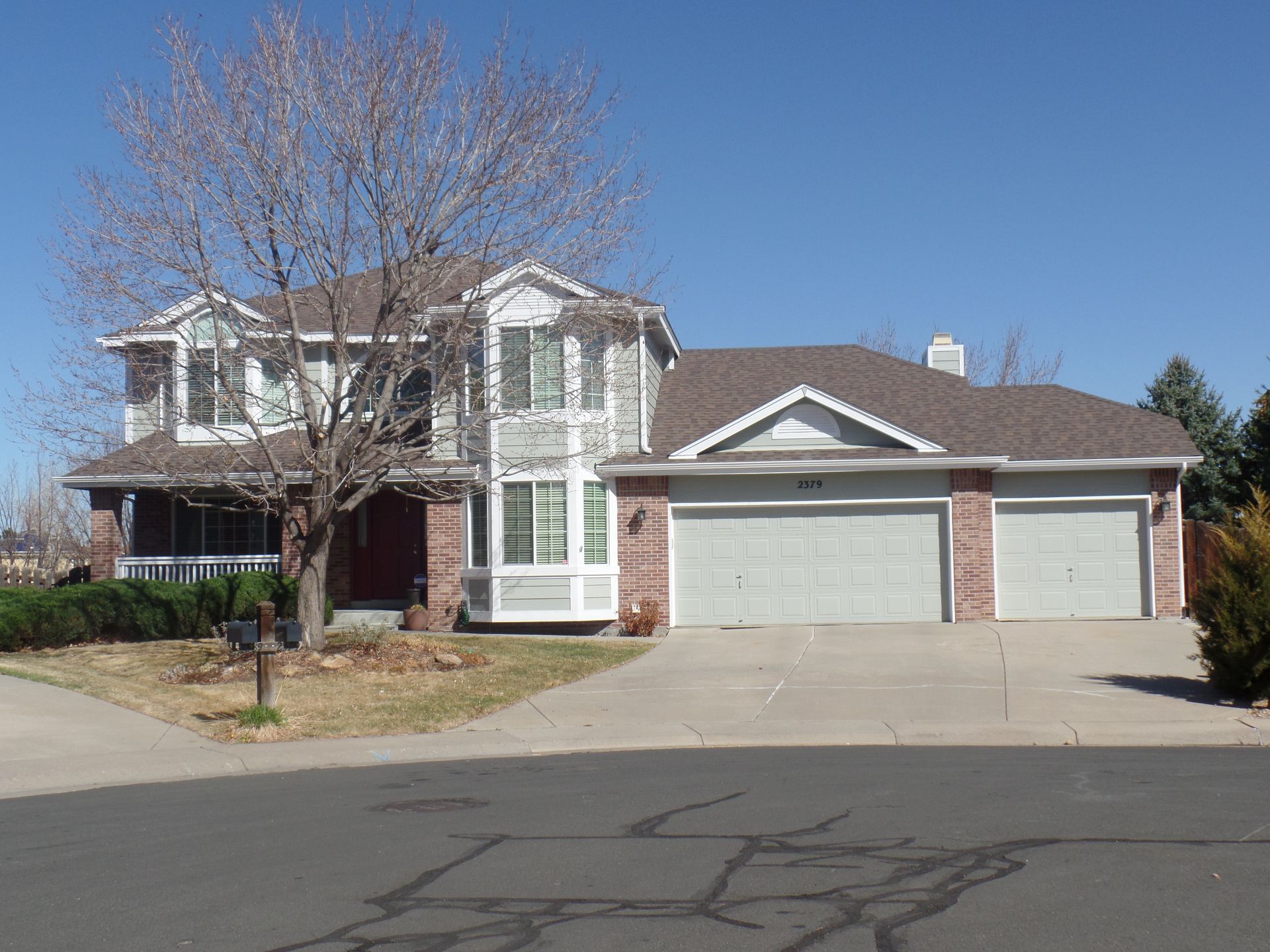 A two-story suburban house with brick and siding, a large tree, a front porch, and a three-car garage.