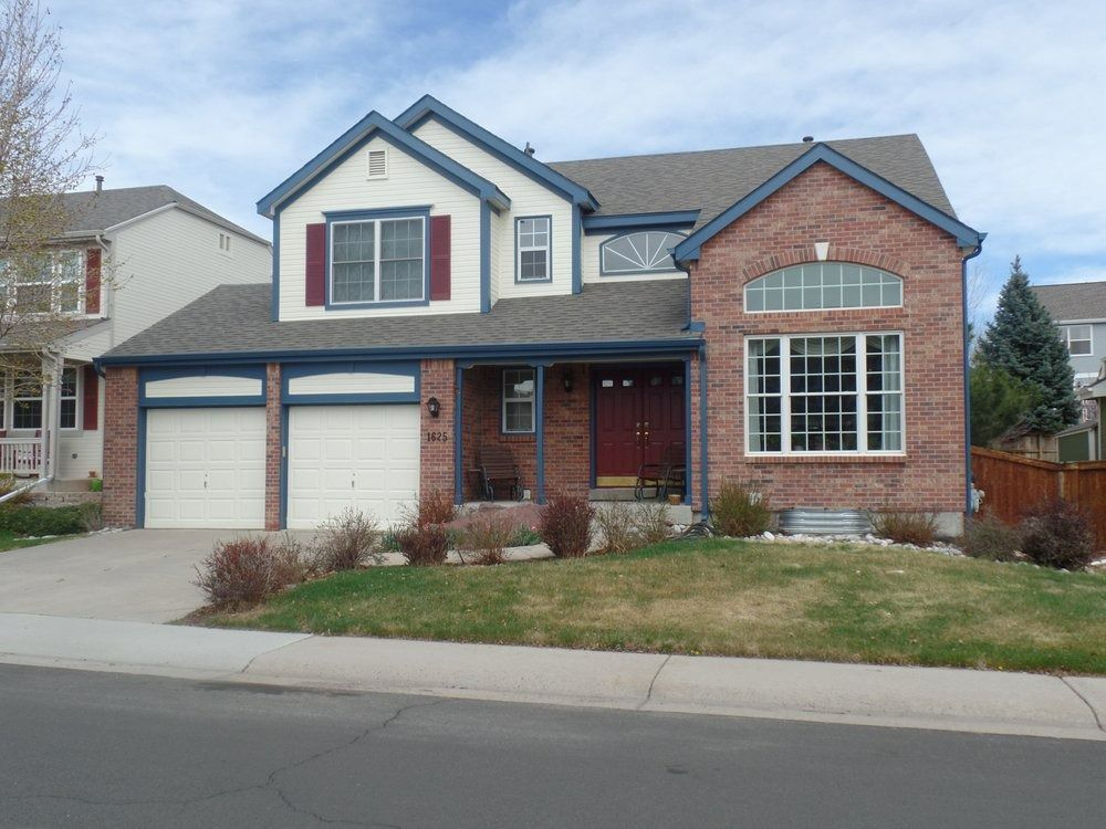 Two-story suburban house with a red brick facade, white trim, red shutters, and an attached two-car garage.