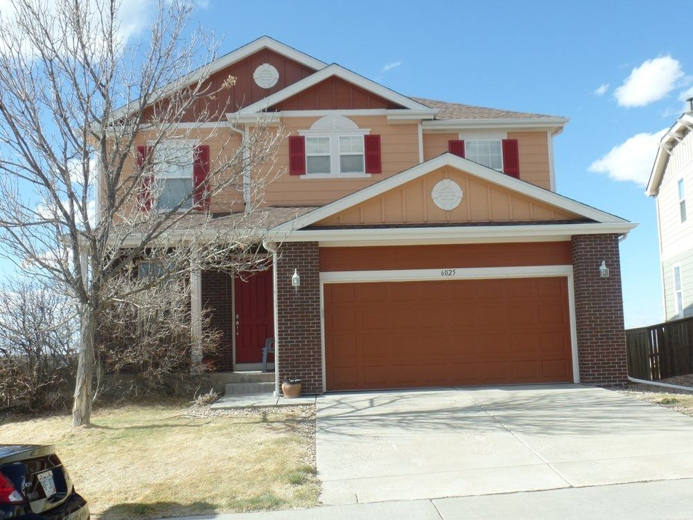 Two-story house with orange exterior, red shutters, and brick accents. Brown garage door and clear blue sky.