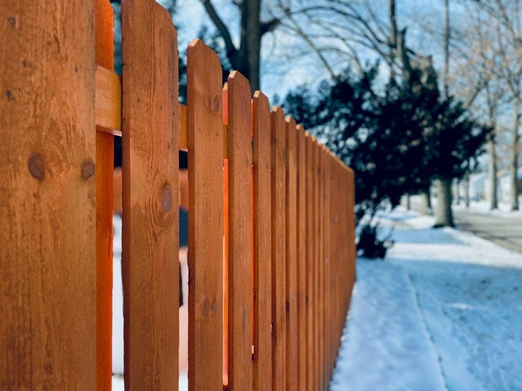 A side view of a tall, light brown wooden picket fence lining a sidewalk covered in patches of snow and bare trees.