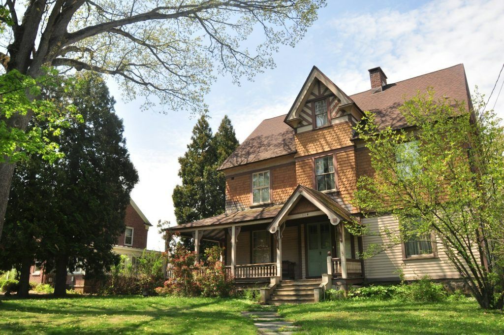 Two-story house with a porch and gabled roof, set on a grassy lawn with trees and a cloudy sky.
