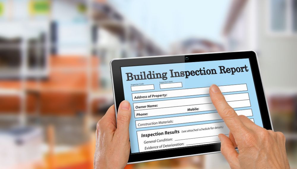 Hands holding a tablet displaying a building inspection report in front of a blurred building interior — Scott MacTaggart Building & Pest Inspections In Old Bar, NSW
