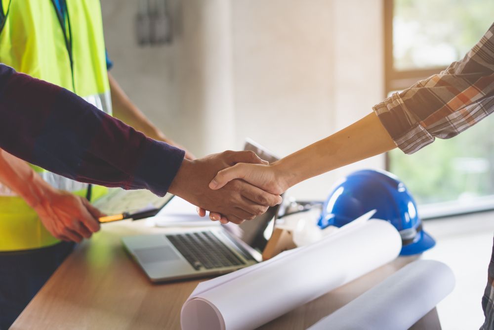 Two people shaking hands over a desk with blueprints, a laptop, and a hard hat; an agreement - — Scott MacTaggart Building & Pest Inspections In Old Bar, NSW