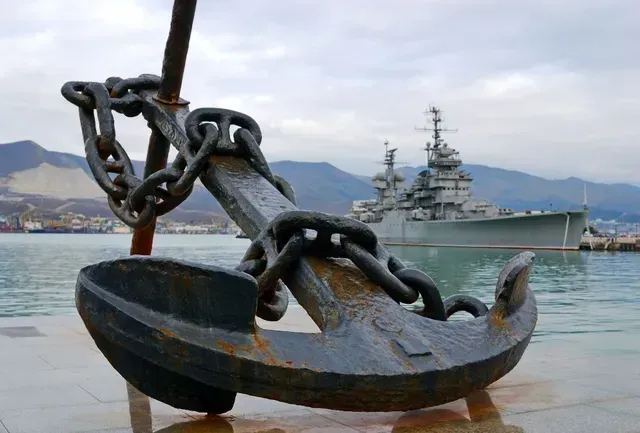 A large anchor on an embankment with a warship in the background