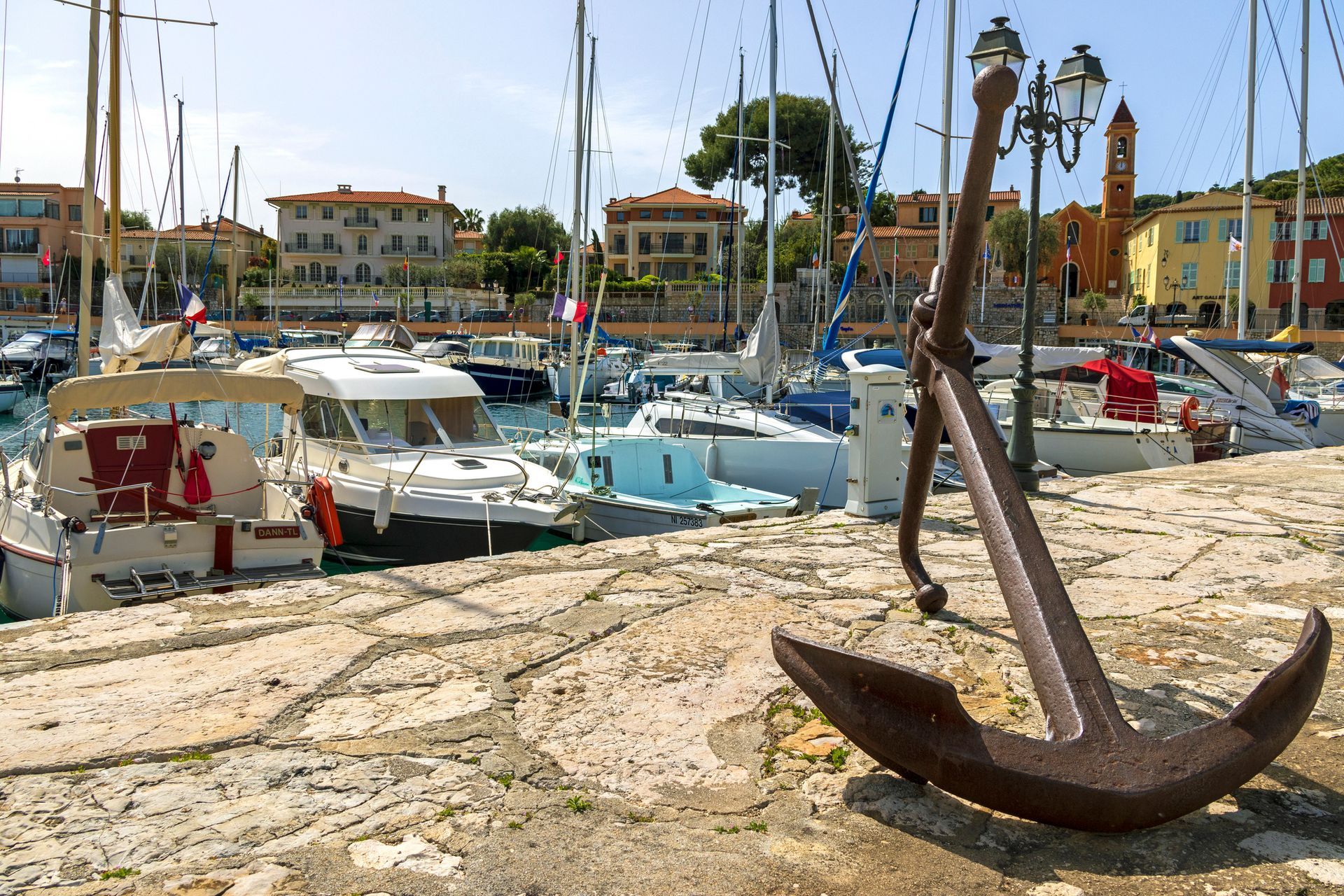 A large anchor is sitting on the ground in front of a marina filled with boats.