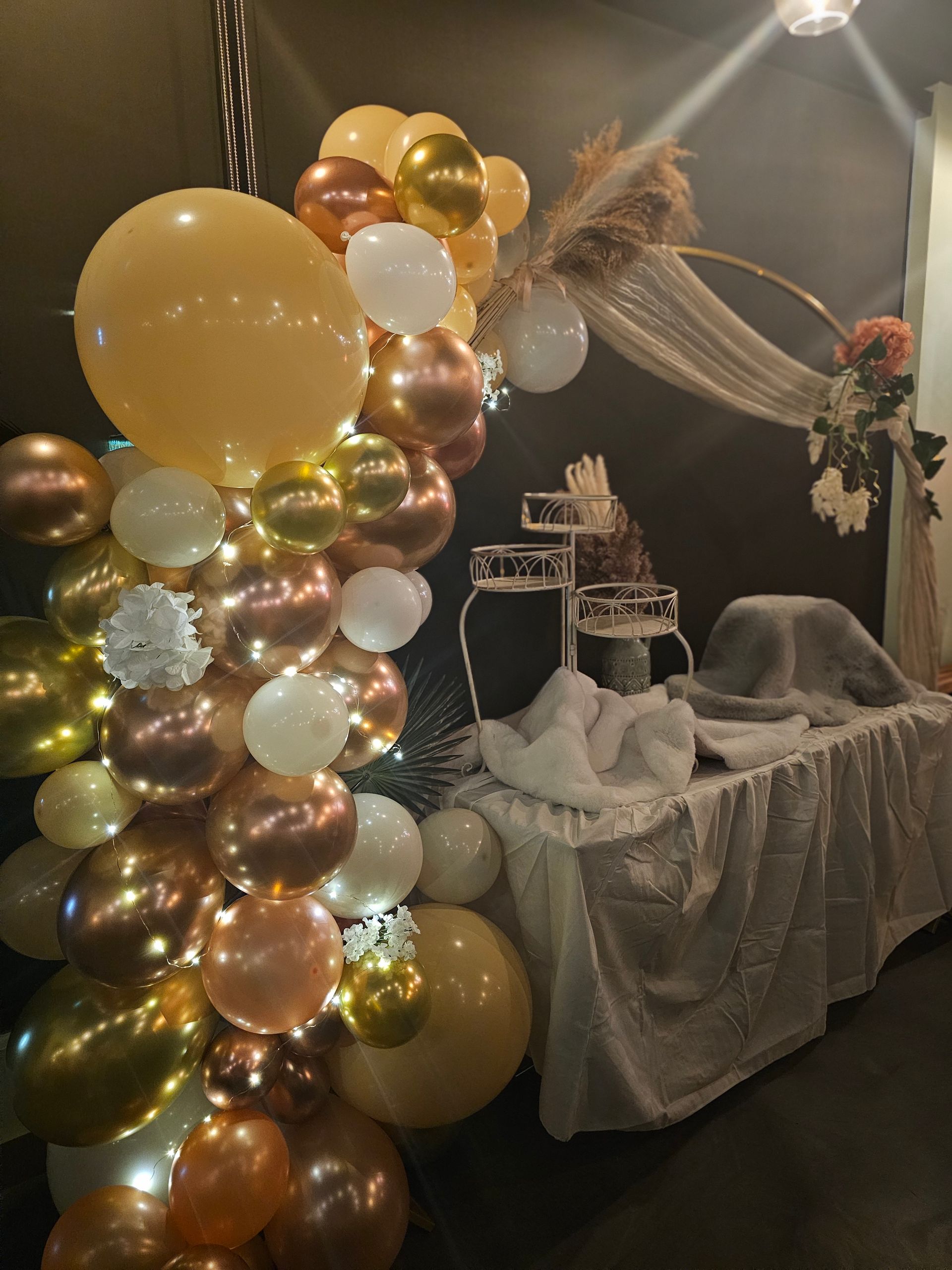 Arche de ballons ornés de ballons dorés, blancs et cuivrés, table de desserts décorée de touches florales.