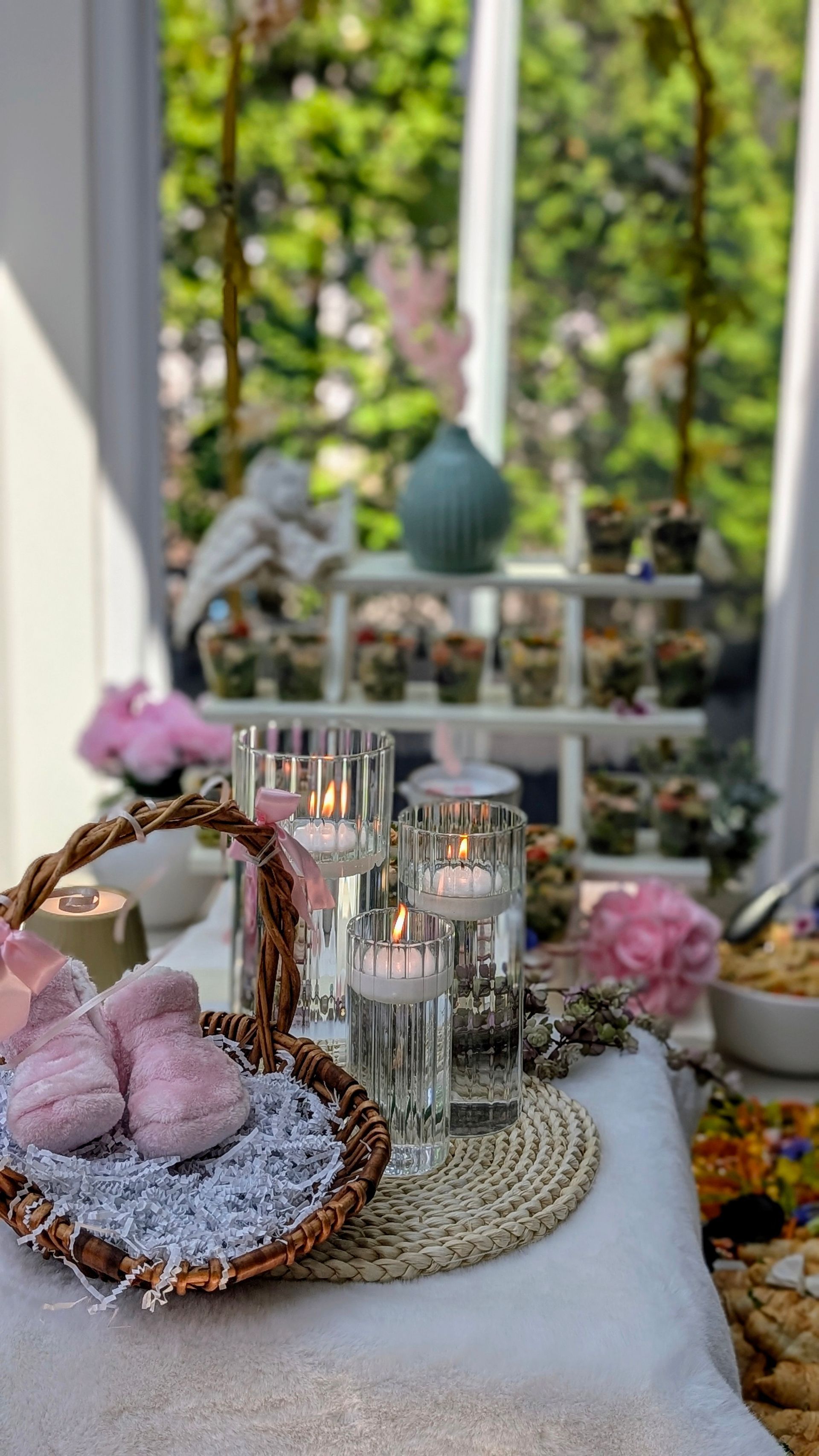 Table de baby shower avec des chaussons roses dans un panier, des bougies flottantes et des desserts, installée à l'extérieur.