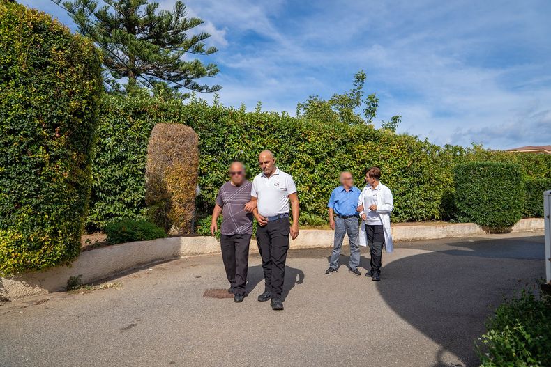 Quattro persone camminano su un sentiero fiancheggiato da siepi; un uomo viene aiutato. All'aperto, soleggiato.
