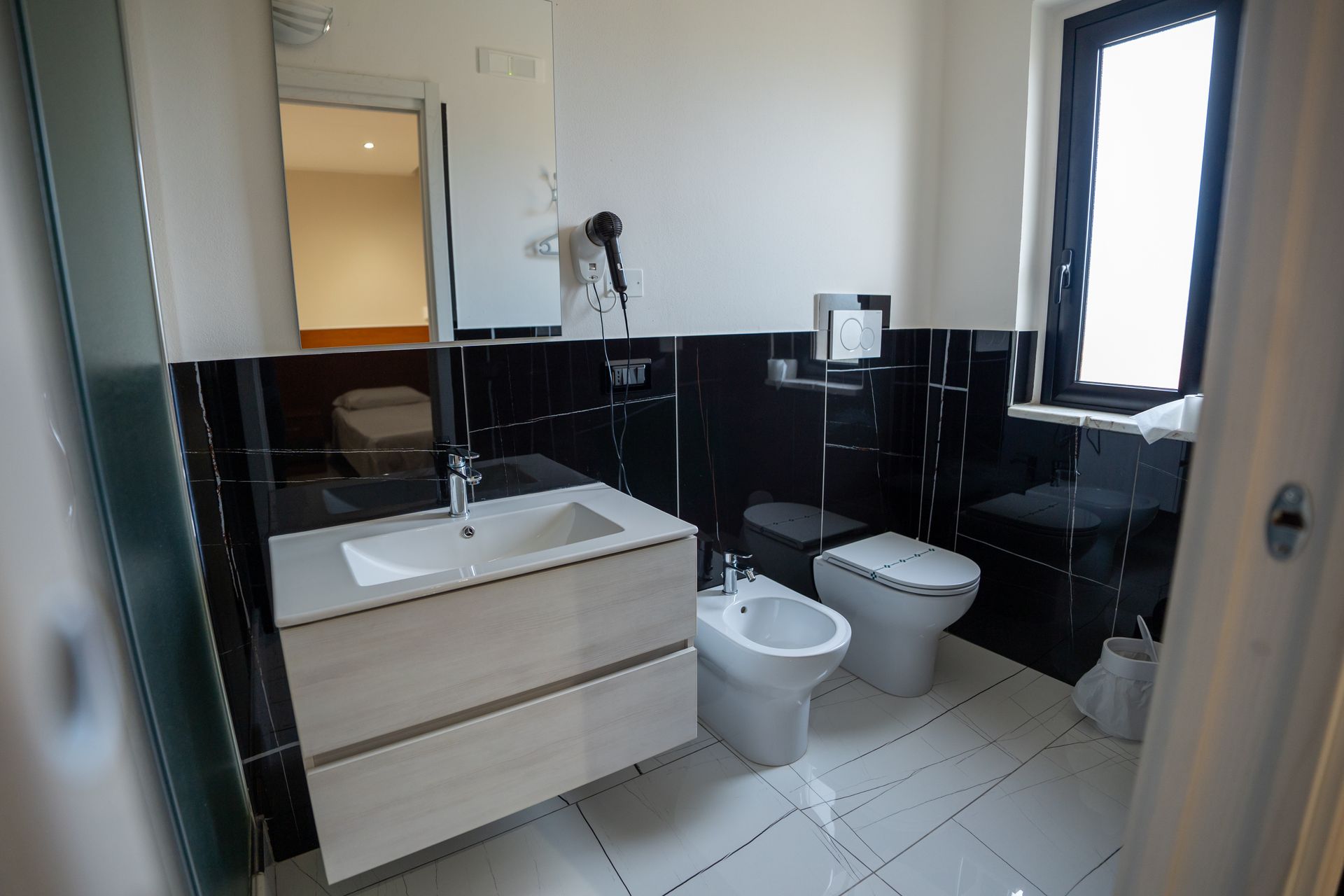 Bagno con piastrelle bianche e nere, lavandino, WC e bidet. Una finestra si trova sul lato destro.