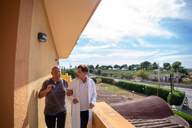 Un uomo e una persona in camice camminano su un balcone con vista sul verde e sulla strada. Giornata di sole.